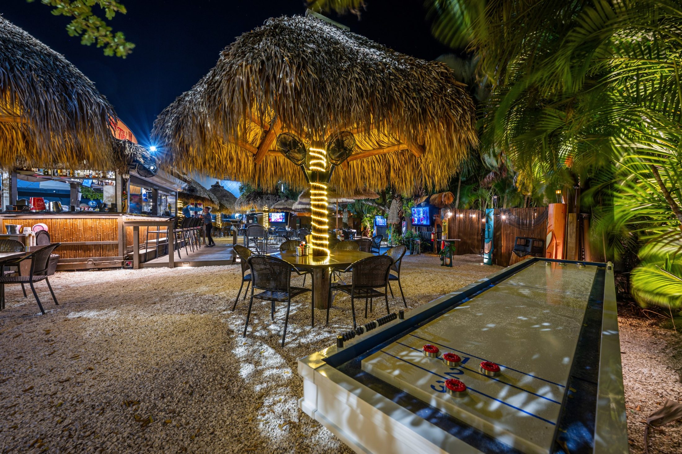 The Cheeky Tiki at night: thatched tiki huts glowing with string lights, a shuffleboard table and palms